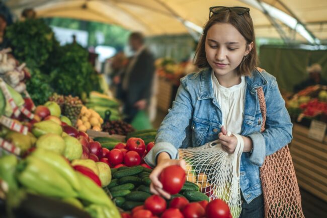 Young girl choosing vegetables and fruit at a farmers market with reusable bag, plastic free and zero waste concept