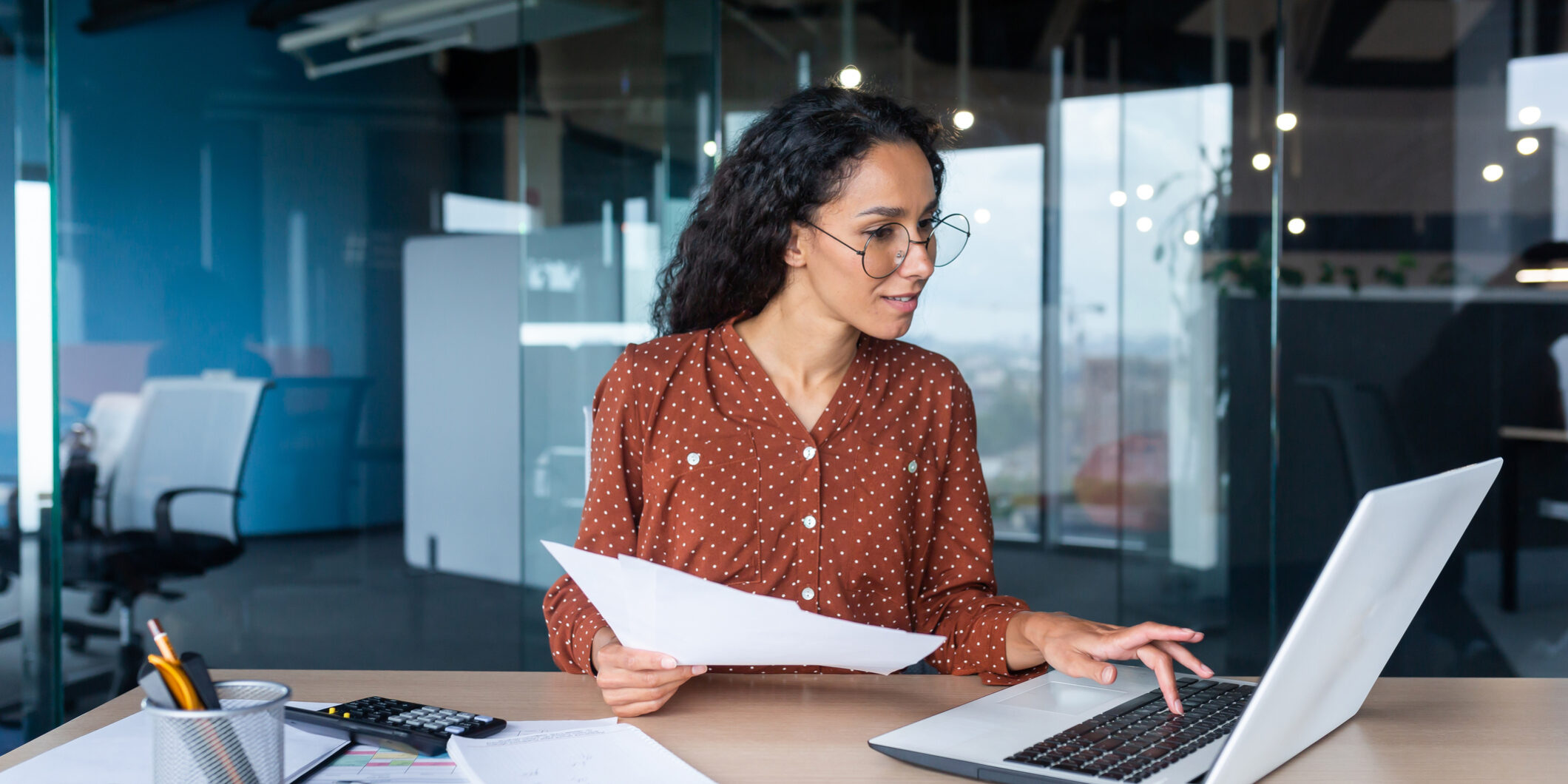 Young happy and successful businesswoman in glasses working with documents inside office, Hispanic woman with laptop looking at bills and contracts, financier with curly hair using laptop