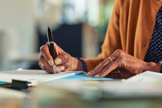African business woman hand writing on paper at desk Close up of african american business woman hands writing on report while working in office. Successful black woman signing documents at home using a pen. Mid adult freelancer signing contract document on table.