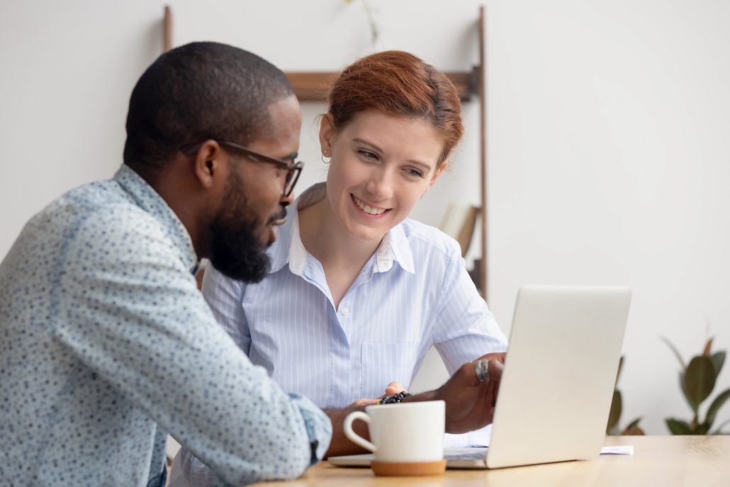 Two diverse smiling businesspeople looking at laptop screen sitting at desk discussing online project, startup, sharing ideas, analyzing results, brainstorming together, thinking of business strategy
