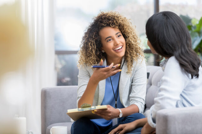 Encouraging therapist talks with young woman Positive young female therapist gestures as she talks with a female client. The therapist smiles warmly as she talks with the young woman.