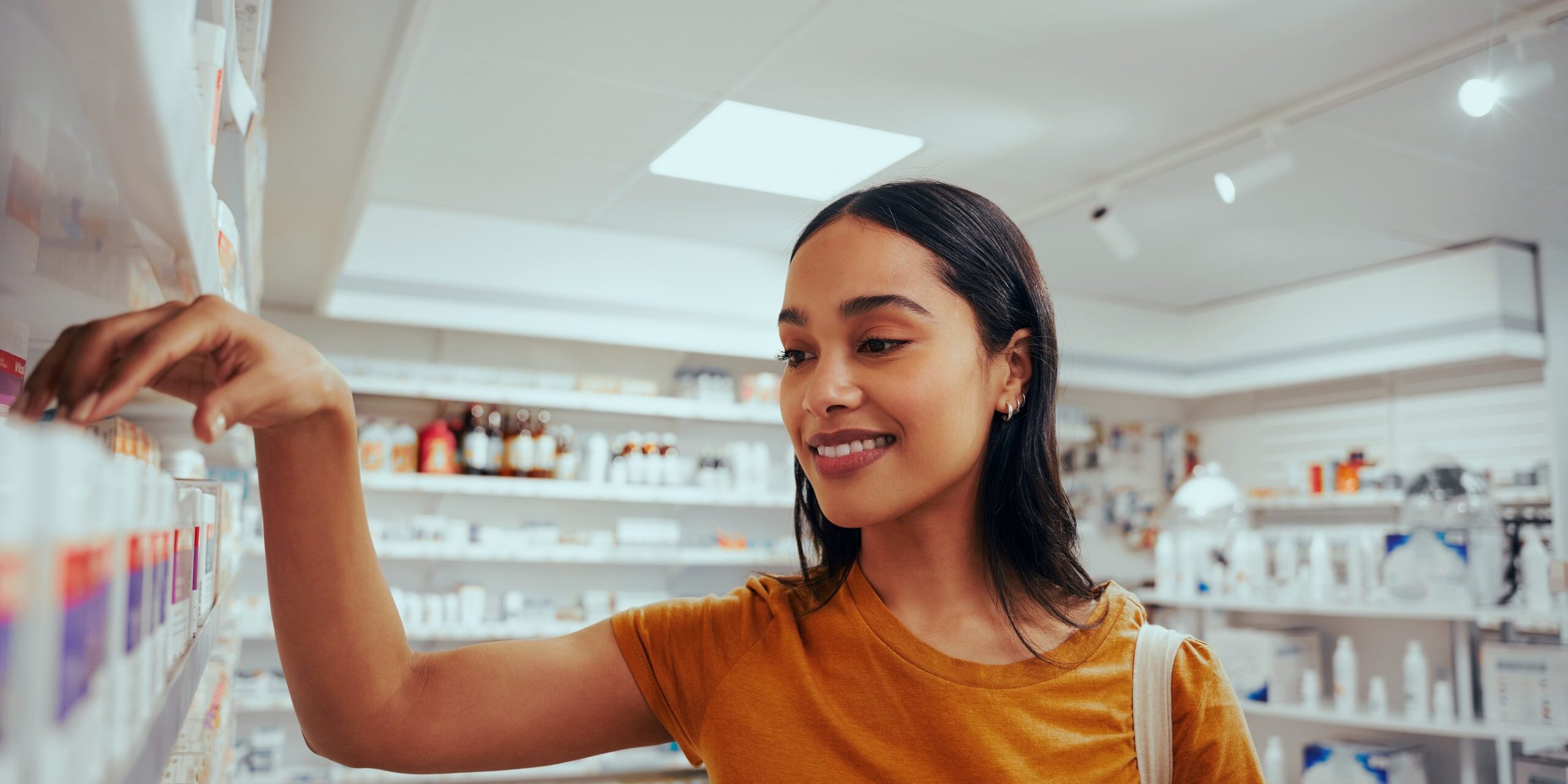 Portrait of smiling young woman in drugstore standing against shelf picking medicine