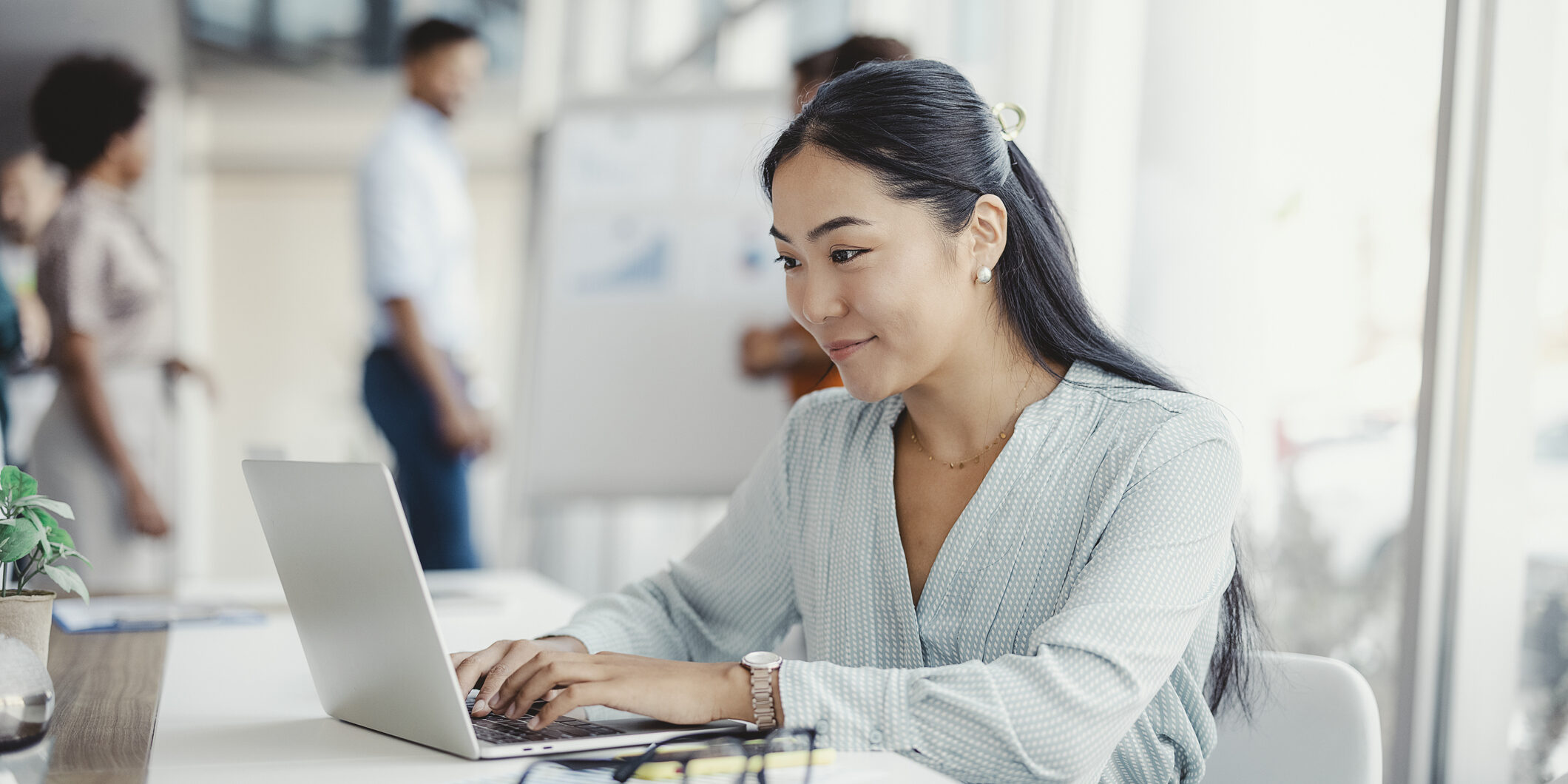 Productivity powered by digital technology Asian woman working on laptop. Businesswoman busy working on laptop computer at office with colleagues in the background.