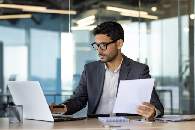 Serious concentrated hispanic businessman financier on paperwork inside office, man in business suit at workplace reviewing and reading papers, contracts and accounts reports.